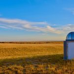 Pequena estação meteorológica instalada em uma extensa pastagem sob céu azul.