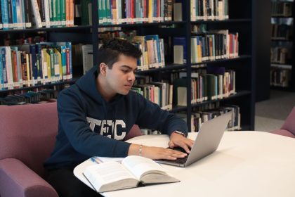 Estudante usa um laptop e consulta livros em uma biblioteca universitária.